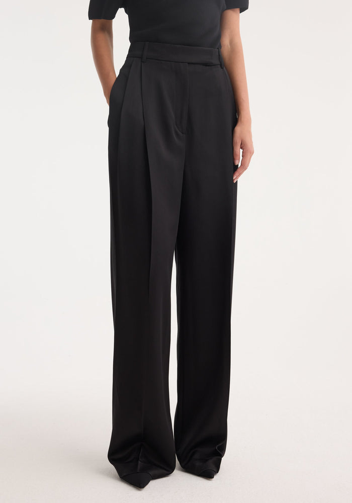 High-waist satin trousers in black, featuring a wide-leg design, front pleats, side pockets, and welt back pockets, modeled to showcase their structured silhouette.