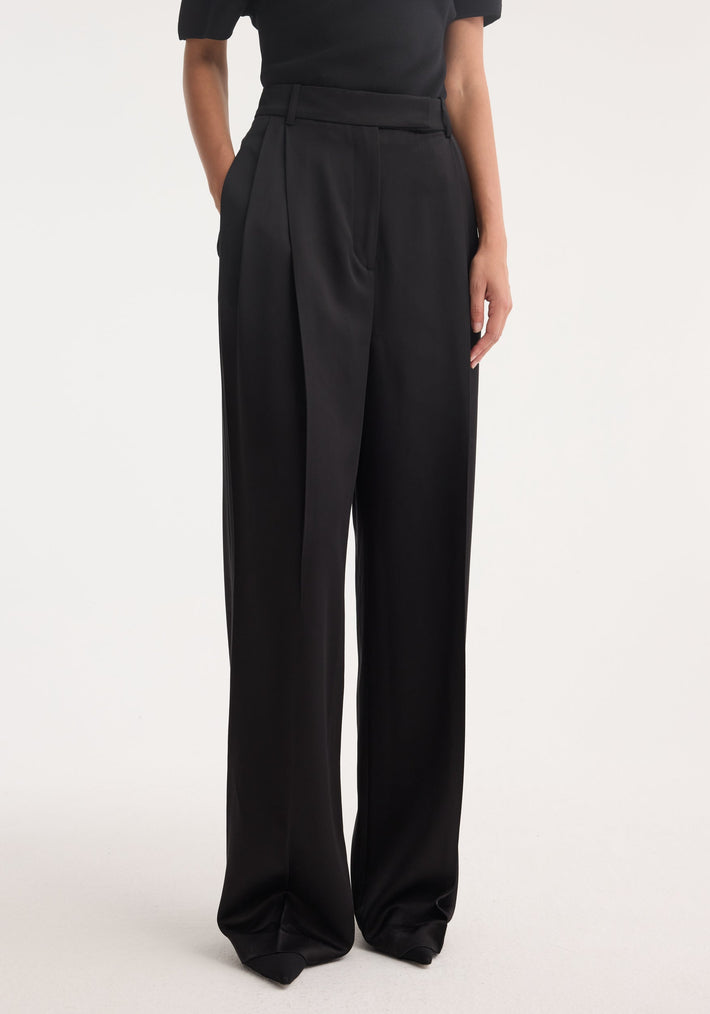 High-waist satin trousers in black, featuring a wide-leg design, front pleats, side pockets, and welt back pockets, modeled to showcase their structured silhouette.