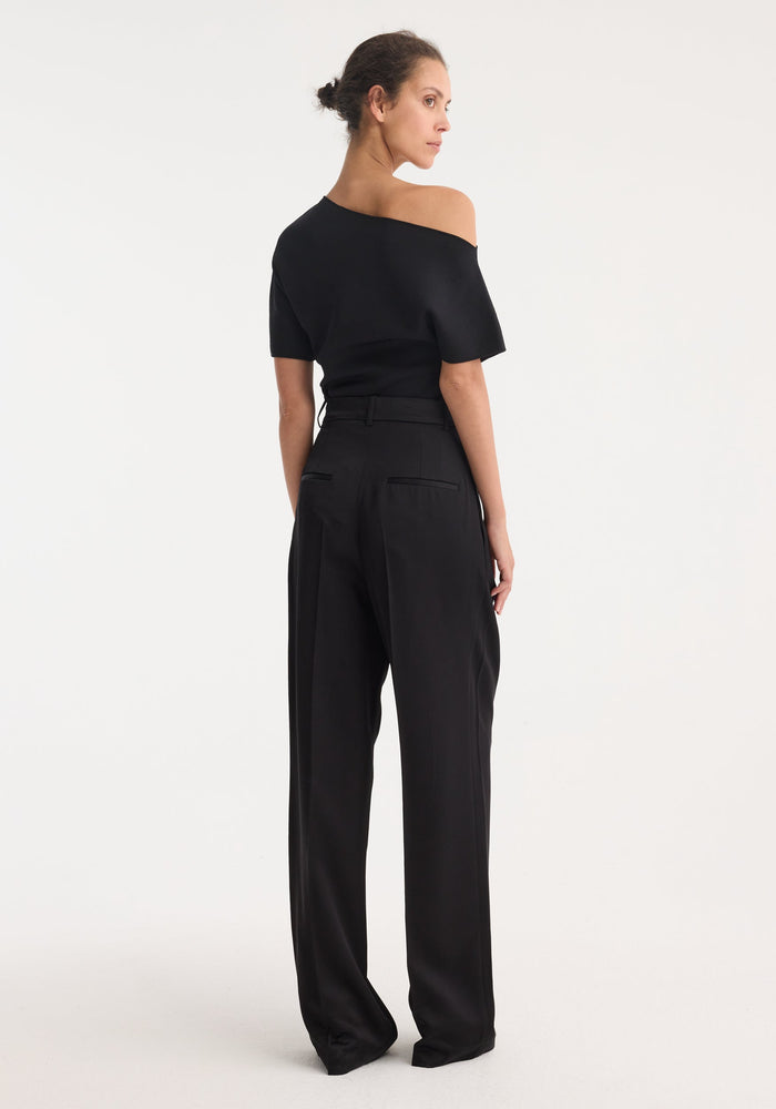 Woman in profile wearing high-waist satin trousers | black, showcasing pleats and welt pockets, highlighting a structured silhouette against a plain studio backdrop.