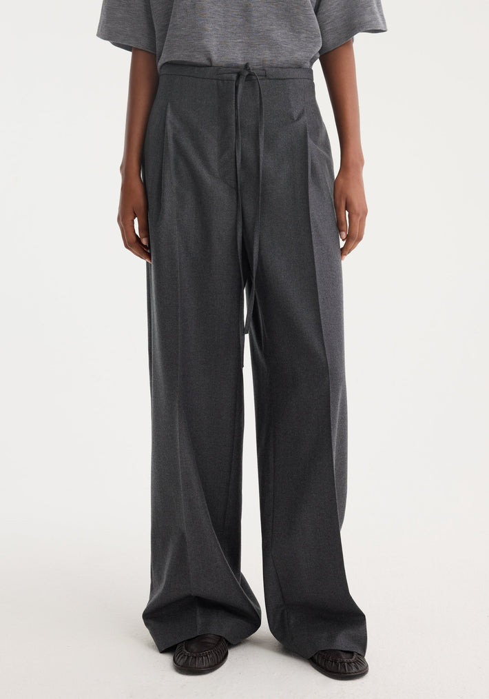 Person wearing Relaxed soft wool trousers, dark grey melange, with wide-leg fit and drawstring waistband, tucked grey top, moccasin slip-ons. Tailored from Italian virgin wool.