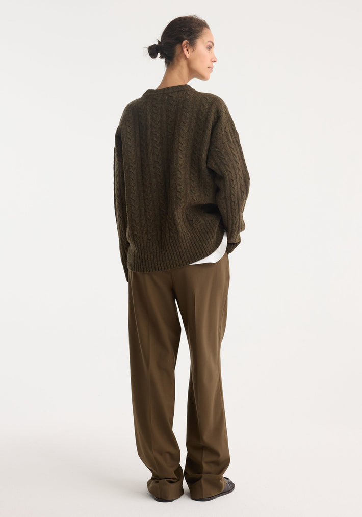 Person wearing an oversized Cable knit jumper | forest melange, featuring a round neckline and dropped shoulders, paired with brown trousers and dark loafers.
