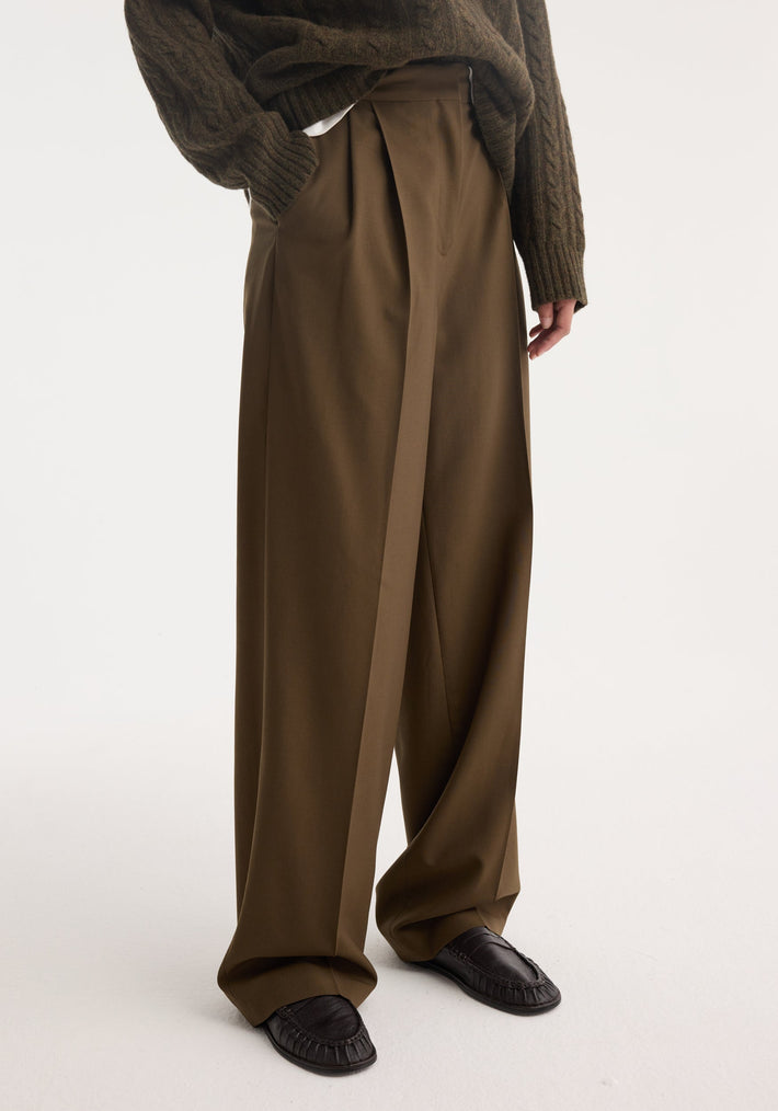 Person wearing Signature tailored trousers in forest hue, featuring wide legs and front pleats, paired with moccasin-style shoes and chunky sweater, showcasing a relaxed silhouette.