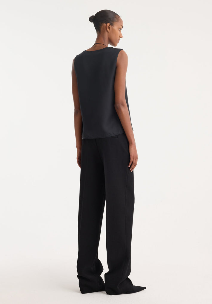 Model wearing high-waisted, straight leg trousers in black, featuring sharp front pleat and tailored waistband. Made from virgin wool blend, showcasing clean side pockets.