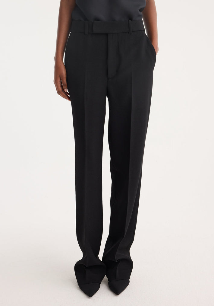 Straight leg trousers in black, featuring high-waisted, tailored fit with sharp front pleat, side pockets, and matching belt, worn over pointed-toe shoes.