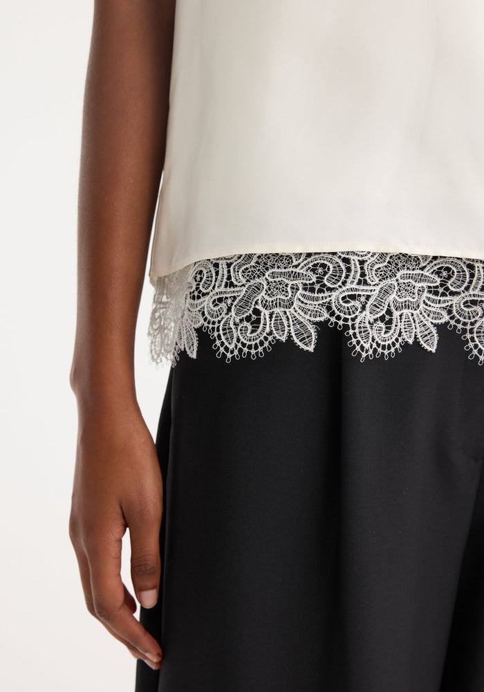 Lace-trimmed silk top in cream with a scoop neck and loose fit, featuring intricate lace detailing on the hem. Model's arm relaxed by their side.
