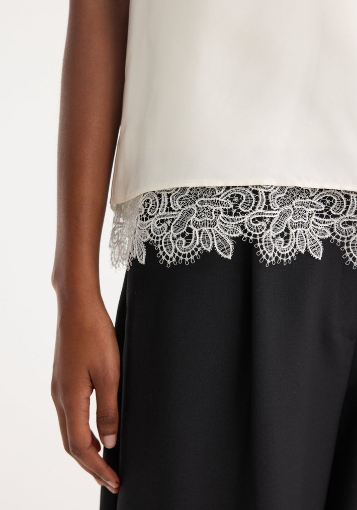 Lace-trimmed silk top in cream with a scoop neck and loose fit, featuring intricate lace detailing on the hem. Model's arm relaxed by their side.