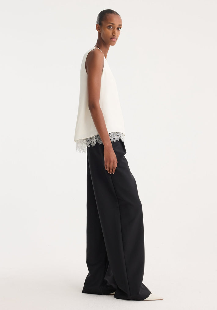 Model wearing a cream lace-trimmed silk top with a scoop neck and loose fit, paired with black wide-leg trousers and pointed white shoes.