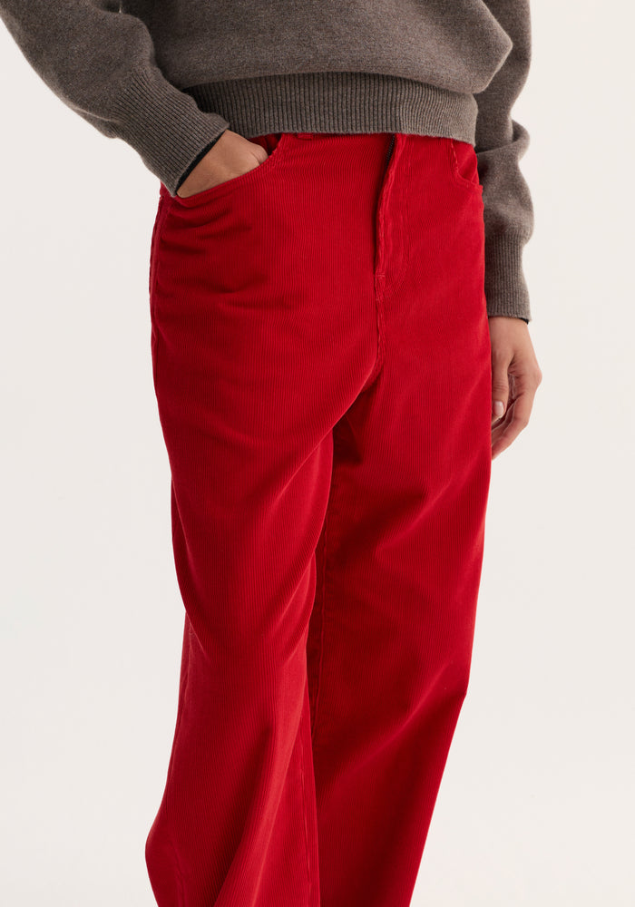Model wearing Relaxed corduroy trousers | rosso with a grey knit sweater; hand in front pocket showing the vibrant red corduroy texture and denim-inspired details.