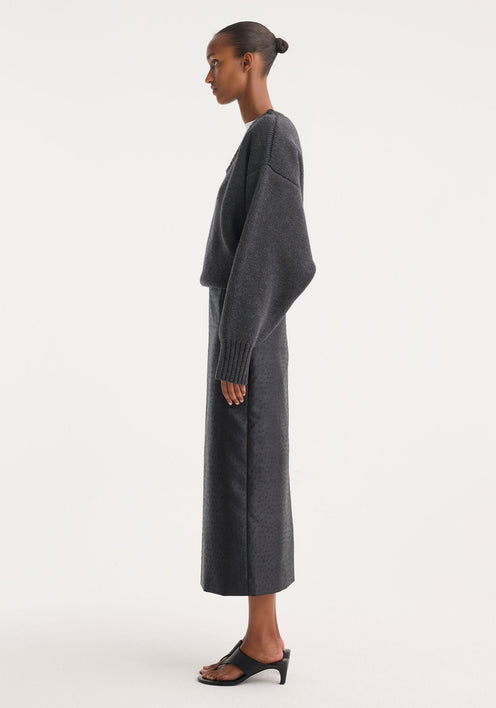 Model in Ostrich satin column skirt, showcasing its high-waisted, embossed texture, paired with a chunky knit sweater, red bag, and black sandals.