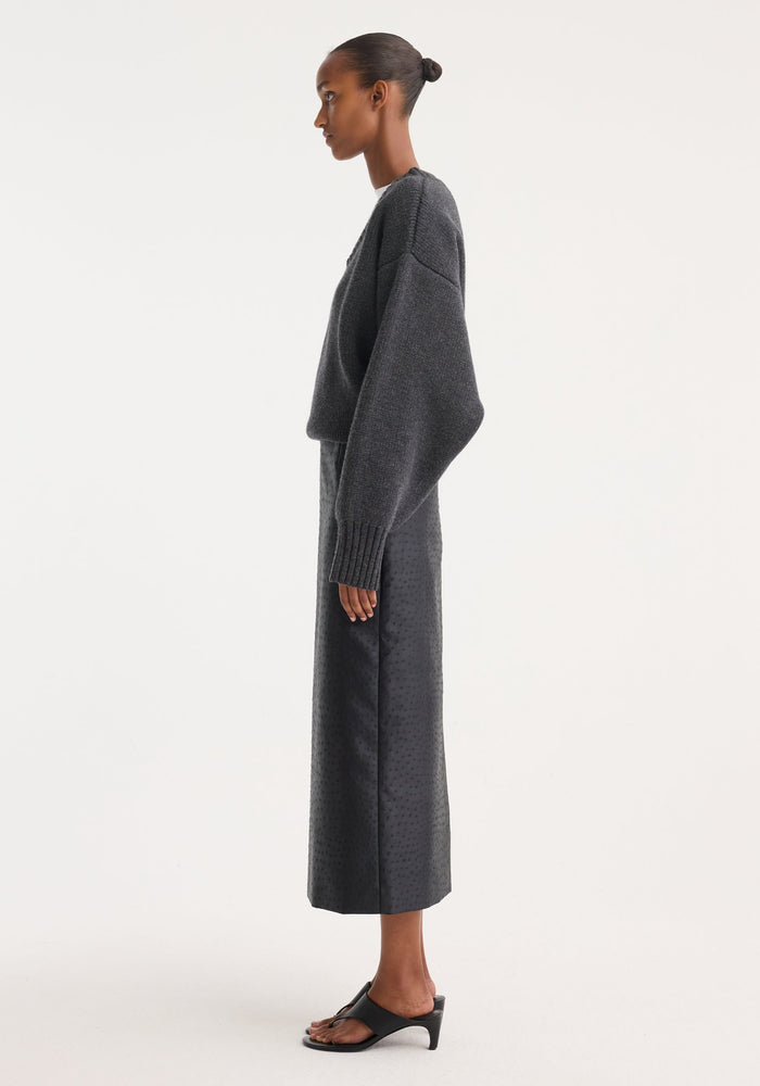 Model wearing Ostrich satin column skirt, showcasing its high-waisted, embossed ostrich-skin texture, paired with a charcoal-gray knit sweater and black heeled slides.