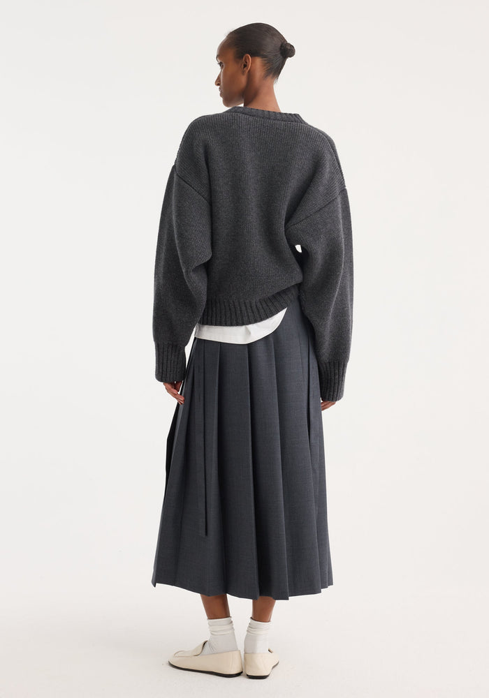 Rear view of a model in a Pleated wrap skirt | dark grey melange, featuring a tailored waistband and voluminous pleats, paired with a loose knit sweater.