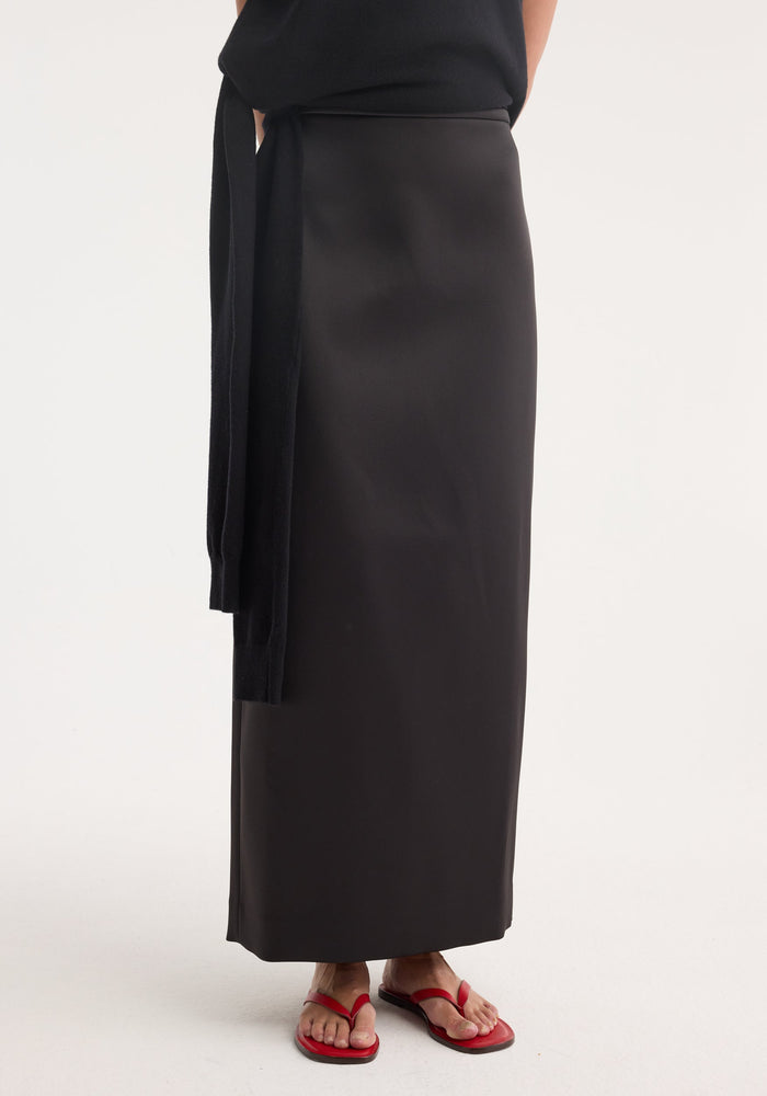 Model wearing Column satin skirt | black, showcasing its high-waisted, ankle-length design, paired with a black top and red sandals against a neutral backdrop.