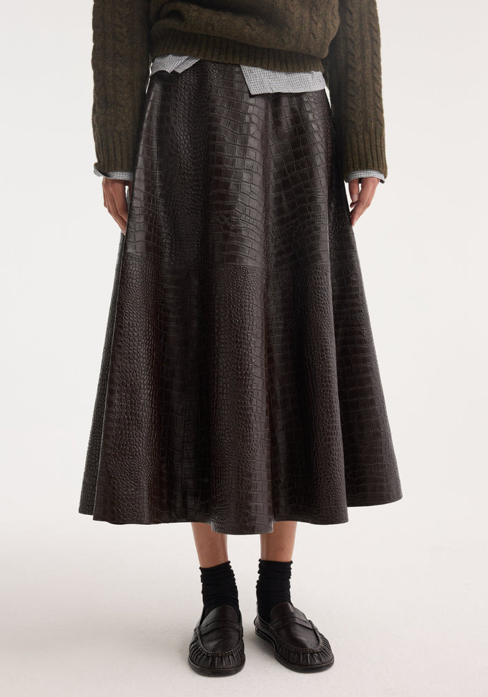 A-line croco skirt in mahogany, featuring a flared silhouette and croco-embossed leather, paired with loafers, cable-knit sweater, and small-check shirt.