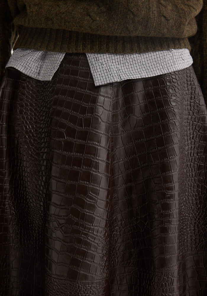 Close-up of A-line croco skirt in mahogany, showcasing its soft, embossed leather texture, layered over a small-check shirt and chunky knit sweater.