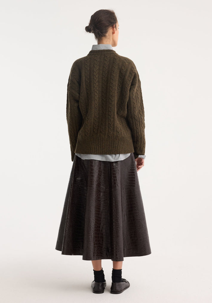 Woman in olive sweater, light shirt, wearing an A-line croco skirt. Mahogany leather panels, embossed pattern, styled with loafers. Model’s back is visible.