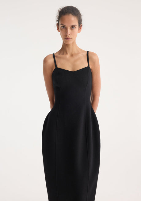 Model wears a Sculptural double-faced dress in black, featuring a bustier-inspired strap top, cinched waist, and mid-calf sculpted skirt with flat sandals.