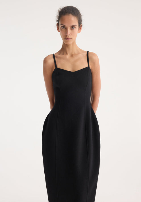 Model wears a Sculptural double-faced dress in black, featuring a bustier-inspired strap top, cinched waist, and mid-calf sculpted skirt with flat sandals.
