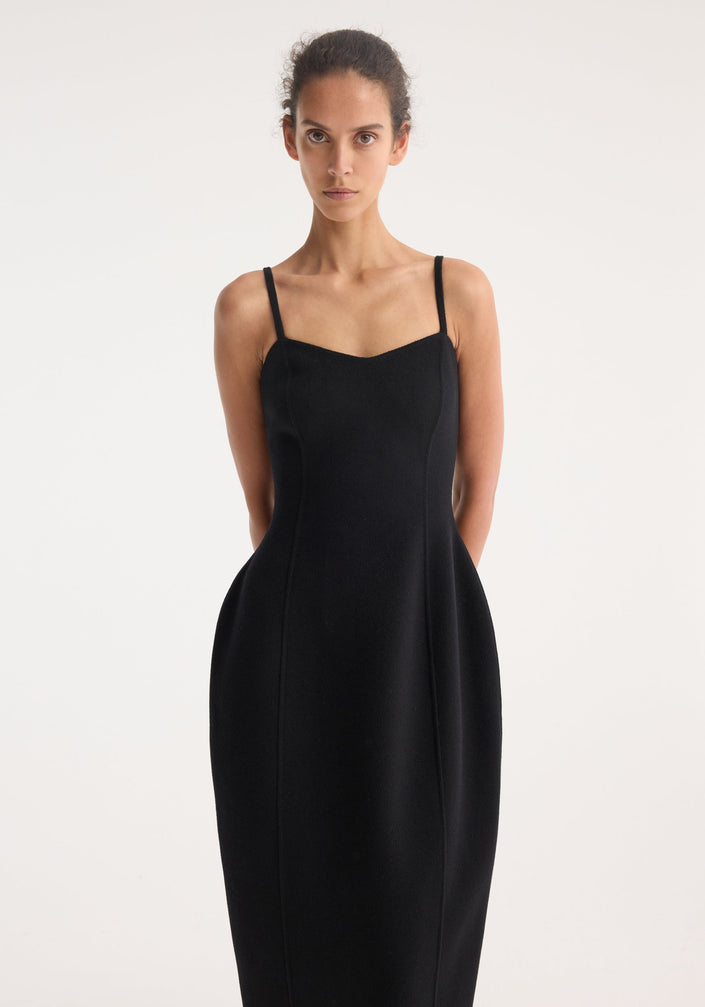 Model wears a Sculptural double-faced dress in black, featuring a bustier-inspired strap top, cinched waist, and mid-calf sculpted skirt with flat sandals.