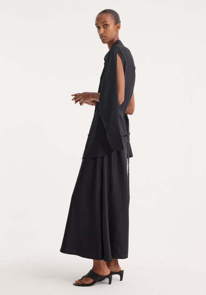 Model wears a Silk gilet-dress with open armholes, featuring a layered, deconstructed design and buttoned wrap belt, creating a fluid silhouette.