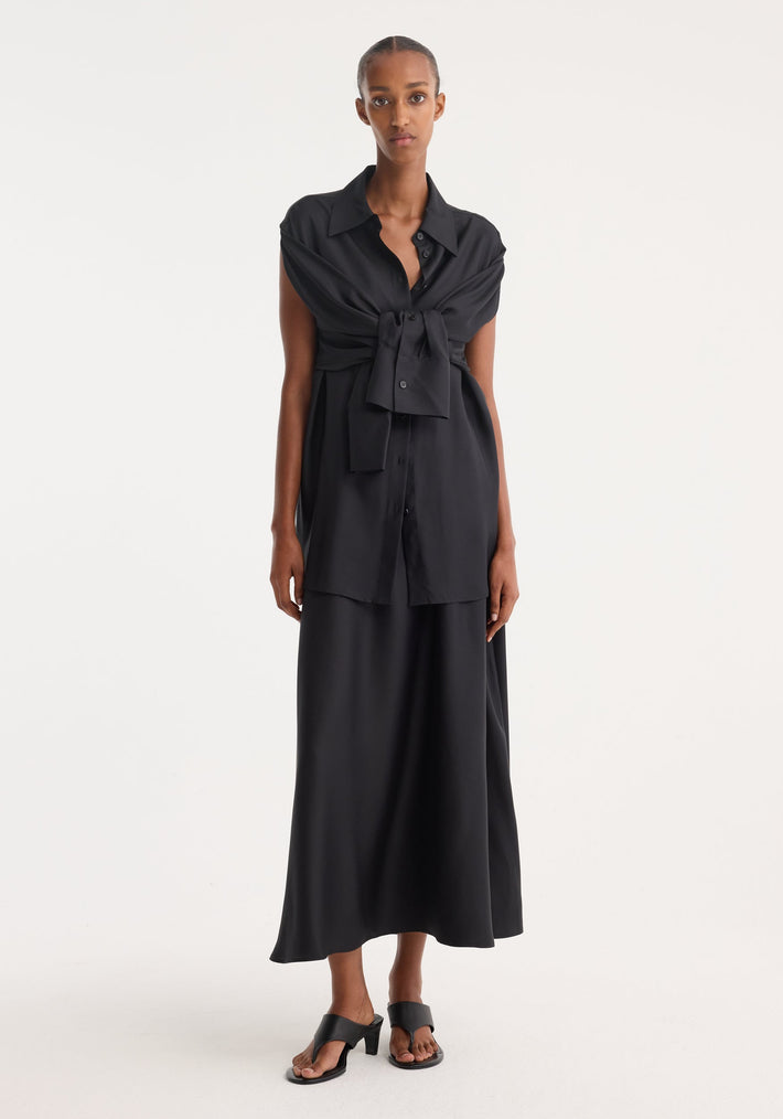 Silk gilet-dress in black, worn by a tall woman; features a flowy skirt, deconstructed shirt with a tie, and black heeled sandals.