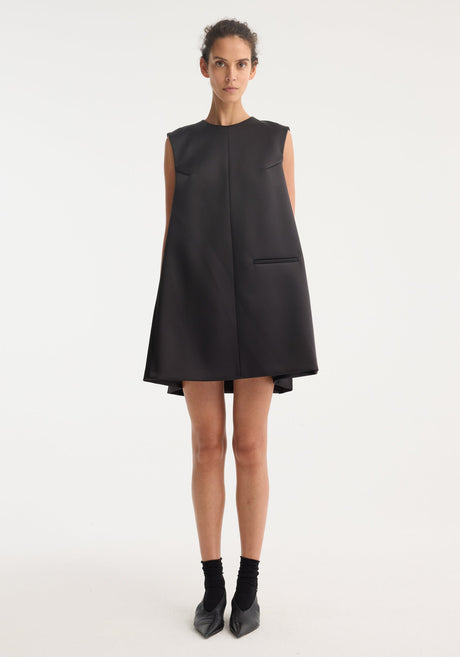 Sculptural cape dress in black, sleeveless A-line with a high neckline, single welt pocket, center-front seam, and dramatic box pleats for a tailored fit.
