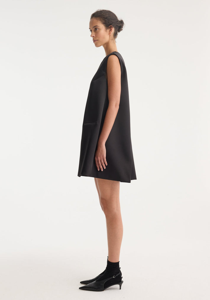 Woman in Sculptural cape dress | black, side profile. Sleeveless, A-line with hip welt pocket, pointed-toe kitten heels, neat bun. Hidden tie for tailored fit.