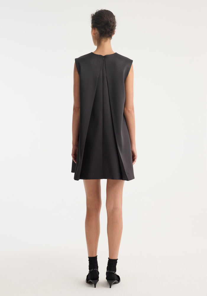 Woman in Sculptural cape dress | black, showcasing a sleeveless A-line mini with dramatic back box pleats, mid-calf socks, and low-heeled Mary-Jane shoes.