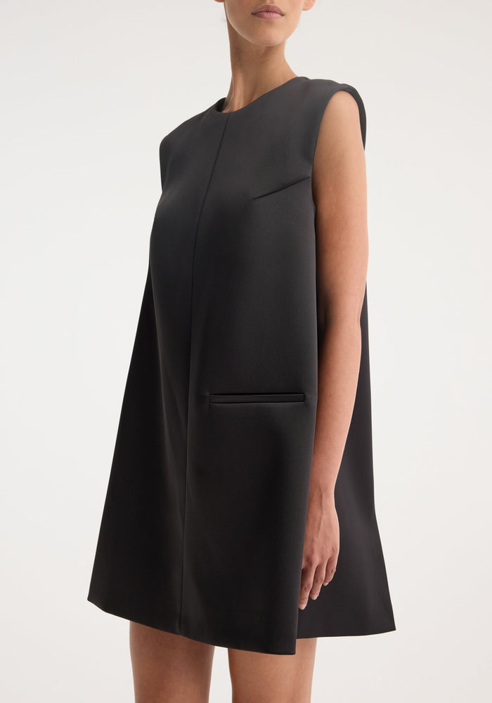 Sculptural cape dress | black: A studio shot of a sleeveless black A-line mini dress with a high neckline and hip-level slit pockets, showcasing minimal seams.