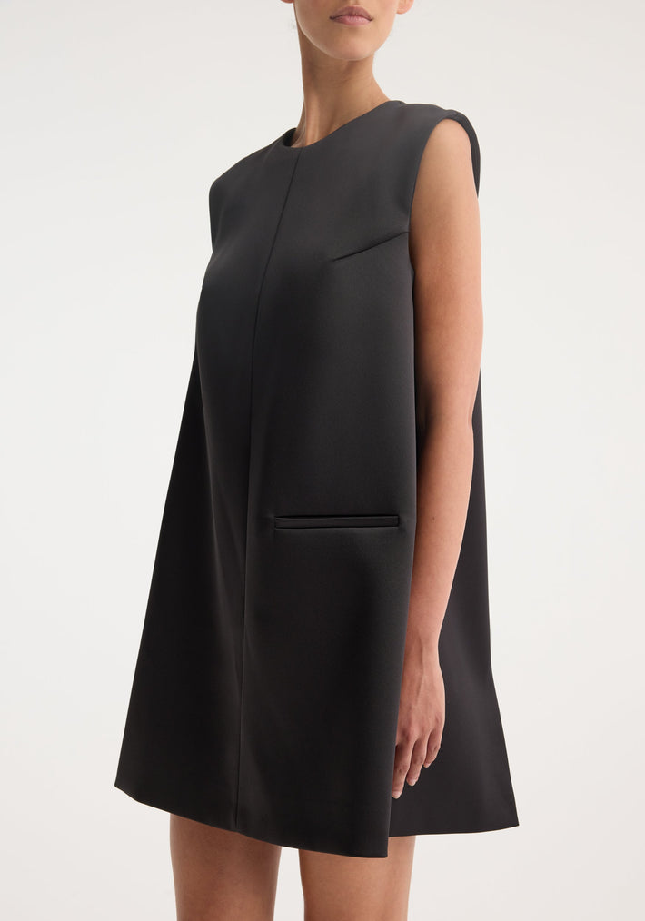 Sculptural cape dress | black: A studio shot of a sleeveless black A-line mini dress with a high neckline and hip-level slit pockets, showcasing minimal seams.