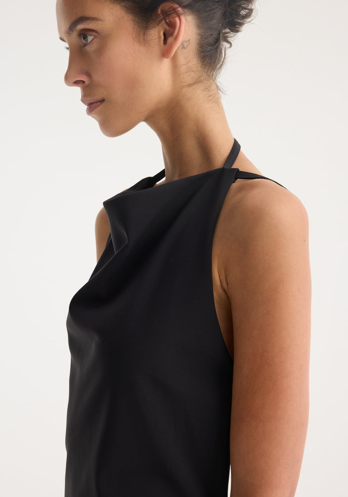 Side-profile of a woman in the Open back dress | black with a draped cowl neckline, thin straps, and visible heart tattoo behind her ear.
