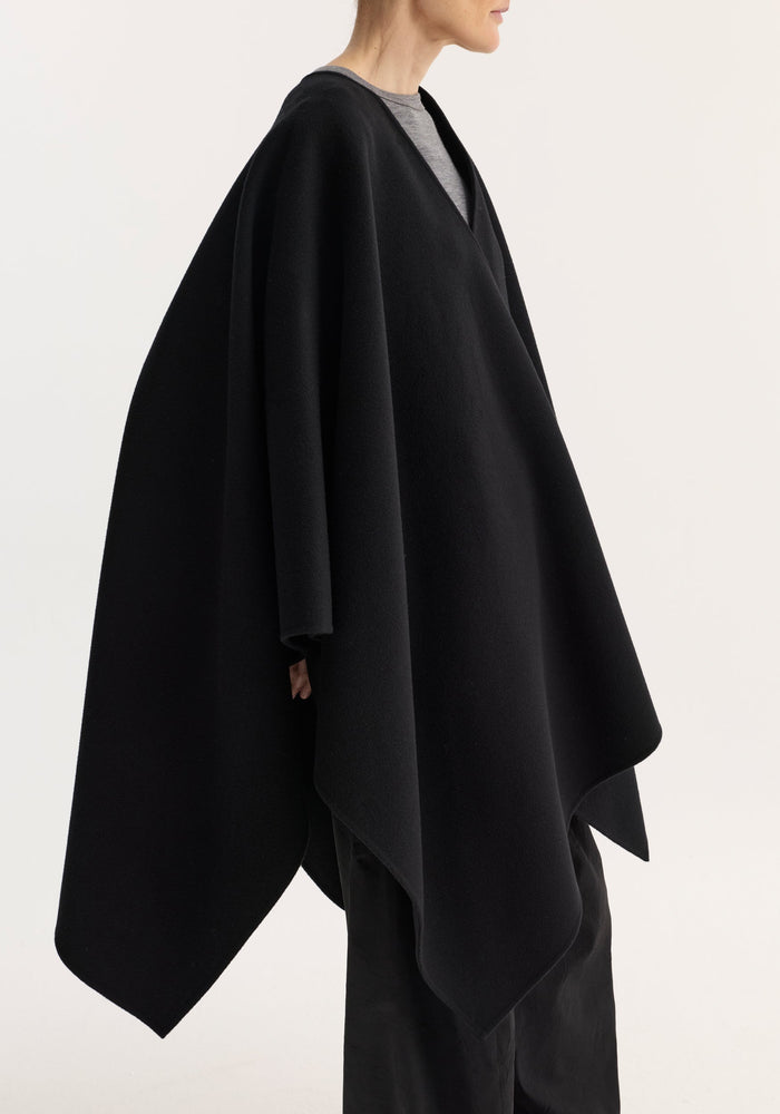 Draped scarf poncho in black, crafted from soft wool, is worn over a grey top with black trousers, showcasing an elegant, asymmetrical design.