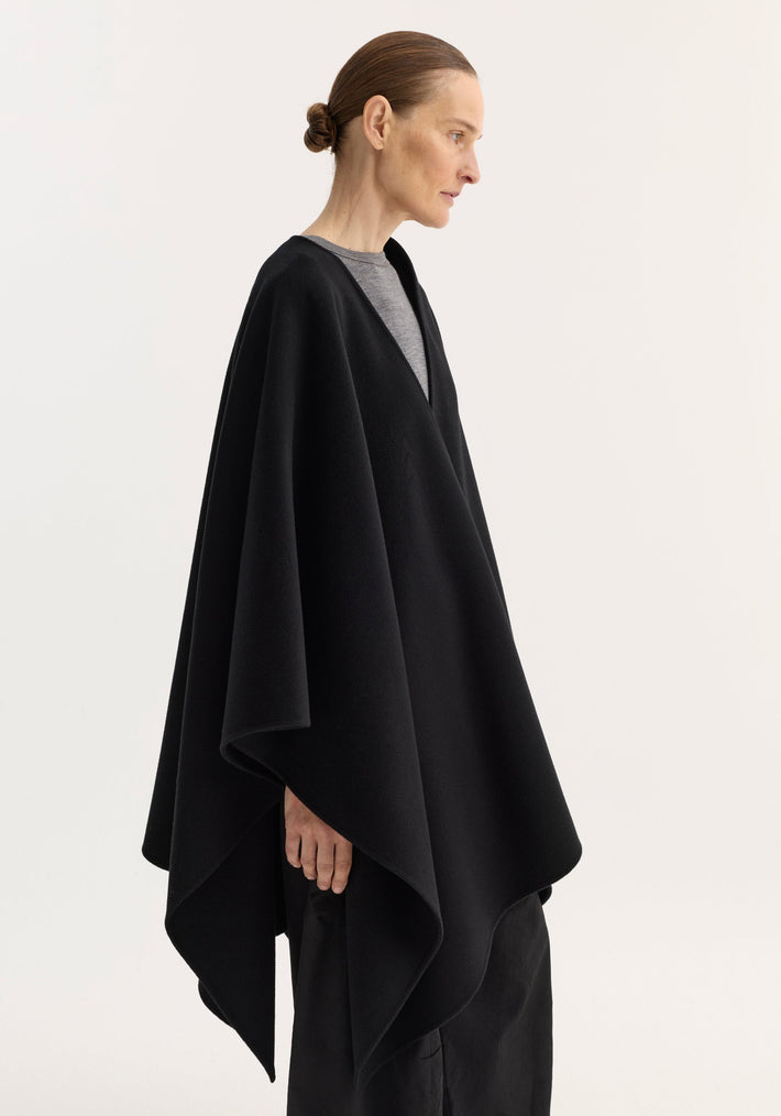 Model in an oversized Draped Scarf Poncho, black, showcasing its asymmetrical hem and soft wool texture, layered over a light top and dark skirt.