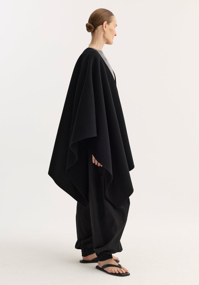 Draped scarf poncho in black wool; voluminous, flowing silhouette on a model, paired with loose trousers and flip-flops, showcasing minimal, monochromatic style.