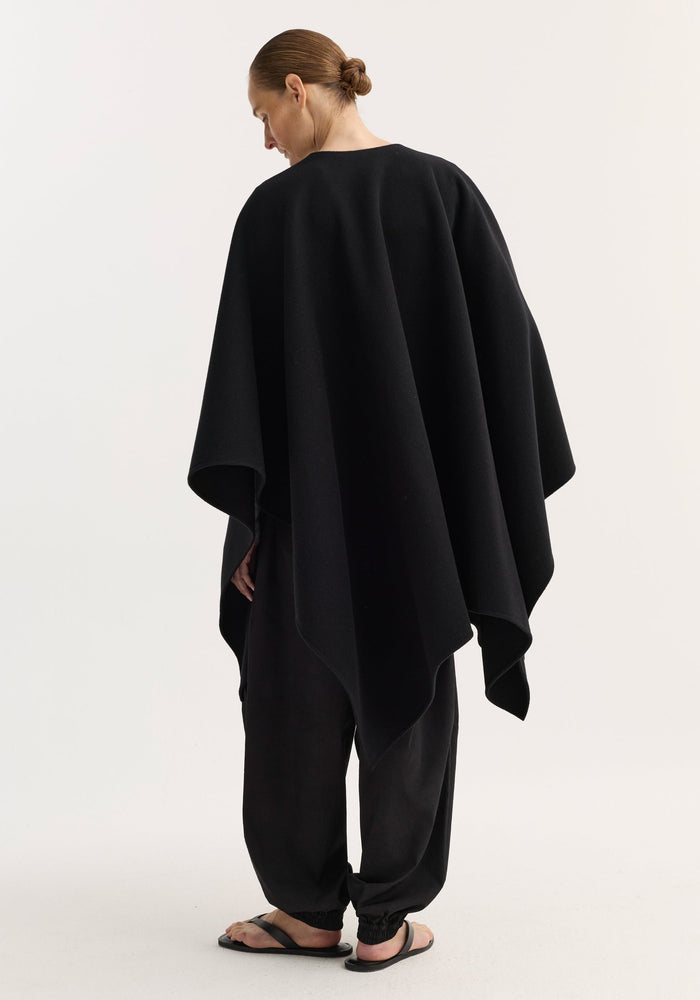 Draped scarf poncho in black, made of soft wool, seen worn from behind. It features a loose, flowing design over relaxed-fit pants and flip-flops.