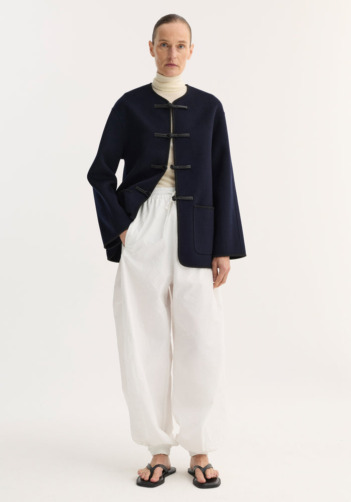 Model wearing Double-faced wool pankou jacket in navy with pankou knot closures, over cream turtleneck, paired with white trousers and black sandals.
