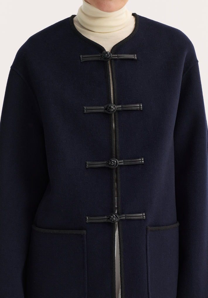 Double-faced wool pankou jacket in navy with black braided trim, round neckline, frog-style toggles, embroidered edges, and patch pockets, worn over a cream turtleneck.