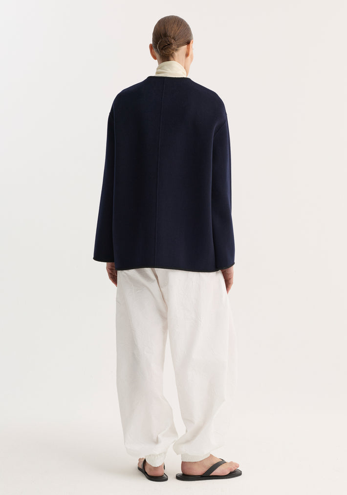 Model wears Double-faced wool pankou jacket in navy, featuring pankou knot closures, embroidered edges, and patch pockets over an ivory turtleneck with white pants.