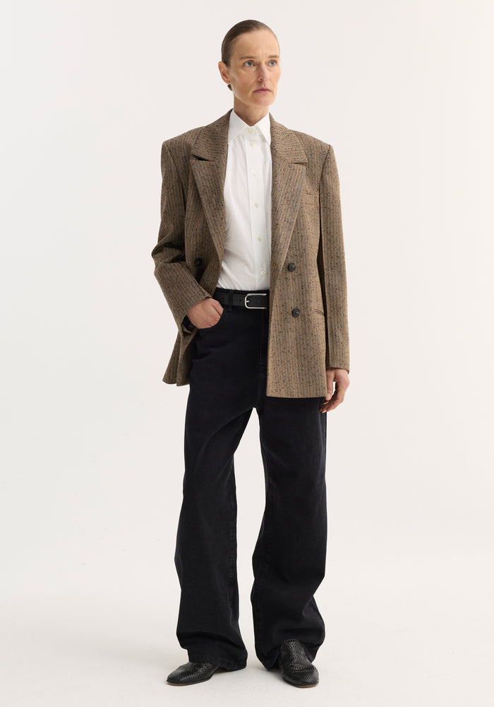 Person wearing a double-breasted blazer with herringbone texture, featuring broad lapels, chest and welt pockets, paired with a white shirt and black trousers.