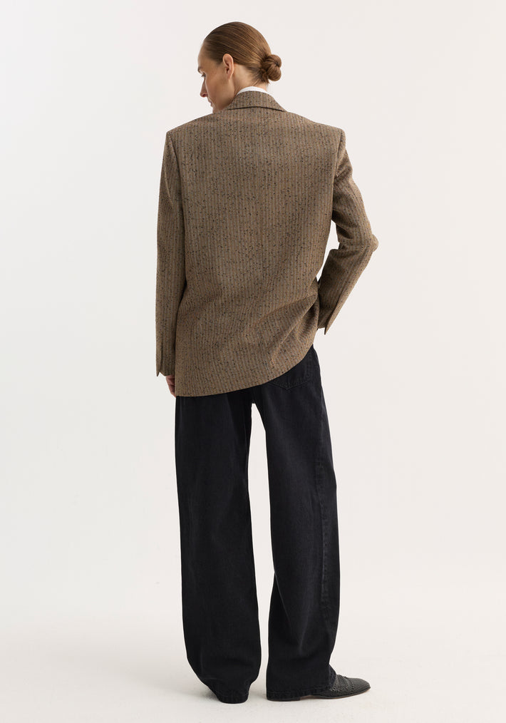 Person wearing a double-breasted blazer in herringbone fabric, featuring clean lines, tailored shoulders, and welt pockets, paired with black trousers and dark woven shoes.