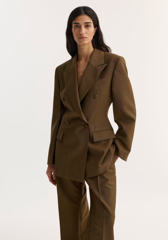 Woman wearing Hourglass blazer, golden brown, with sharp notch lapel, broad shoulders, cinched waist, and matching trousers, hand in pocket, long hair down.