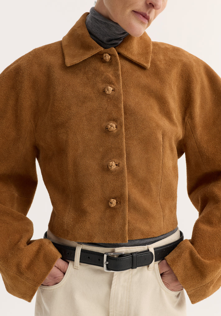Person wearing a Rounded Textured Suede Jacket | Cognac with knotted buttons over a dark turtleneck, hands in pockets of light pants with a black belt.