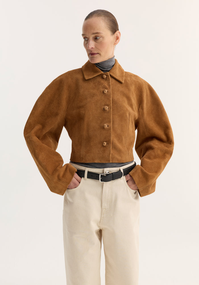 Model wearing the Rounded Textured Suede Jacket in cognac over a grey turtleneck, paired with cream trousers and black belt, showcasing its cropped, structured design.