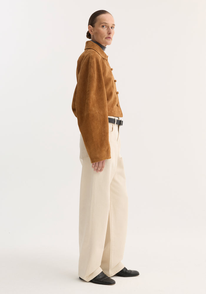 Model in a Rounded textured suede jacket | cognac, showcasing its cropped fit, structured sleeves, and knotted leather buttons, paired with cream trousers and black slip-ons.
