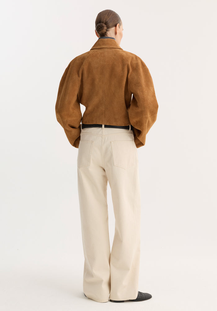 Back view of a model wearing a Rounded textured suede jacket | cognac, featuring a cropped fit, waisted silhouette, and knotted leather buttons, paired with cream trousers.