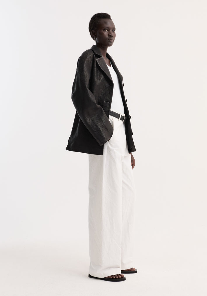 Model in a slightly oversized Leather jacket with small lapel | black, layered over a white tank, paired with high-waisted white trousers and black sandals.