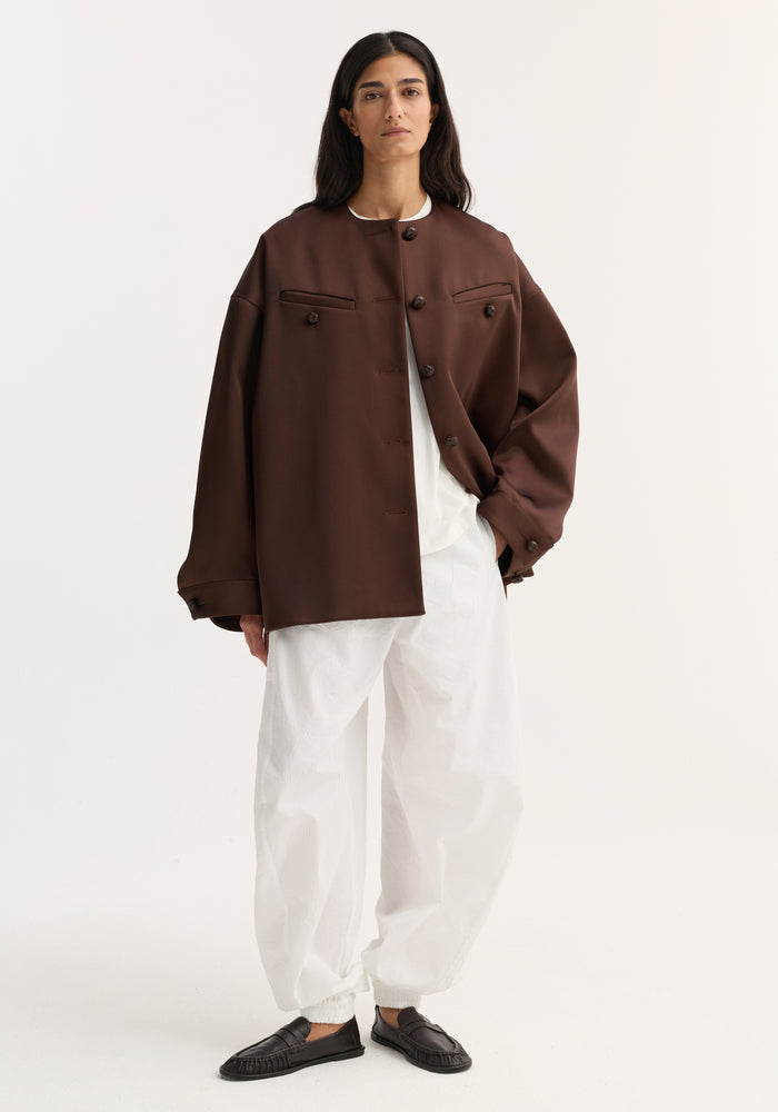 Woman wearing Western heavy satin jacket with leather-knotted buttons, white top and pants, and black loafers against a white background.