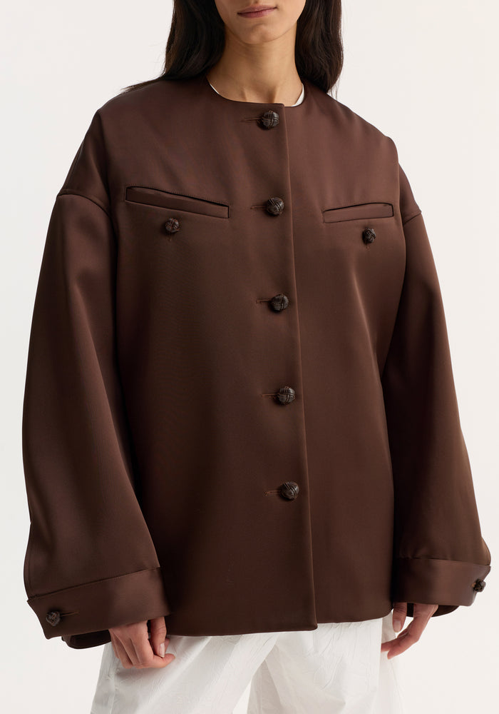Model wearing Western heavy satin jacket | espresso, featuring oversized fit, knot-style buttons, rounded sleeves, and chest welt pockets, paired with white trousers.