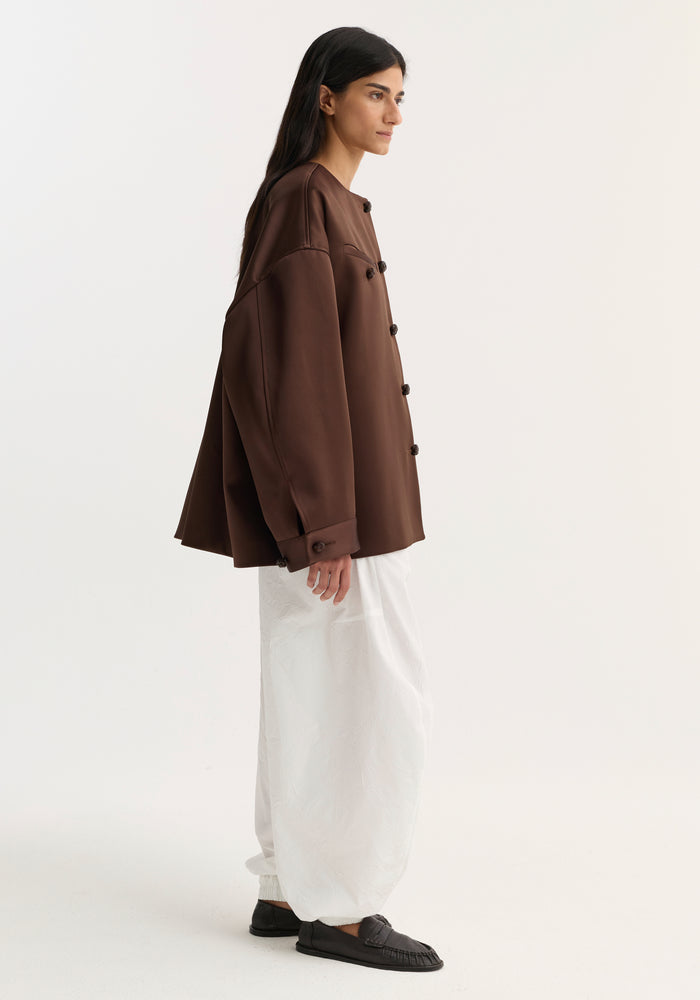 Woman wearing Western heavy satin jacket in espresso, featuring oversized fit, distinctive leather-knotted buttons, and high chest welt pockets, paired with billowy white trousers.
