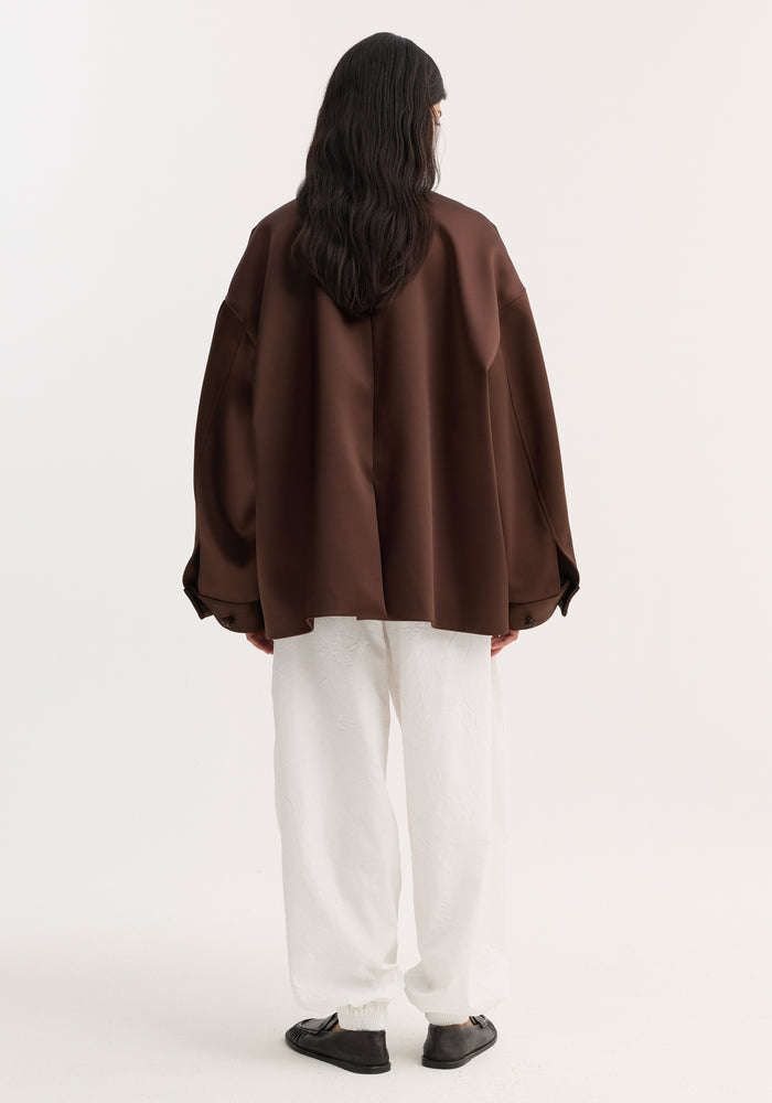 Back view of a model in an oversized Western heavy satin jacket with leather-knotted buttons and chest welt pockets, paired with white tapered trousers.