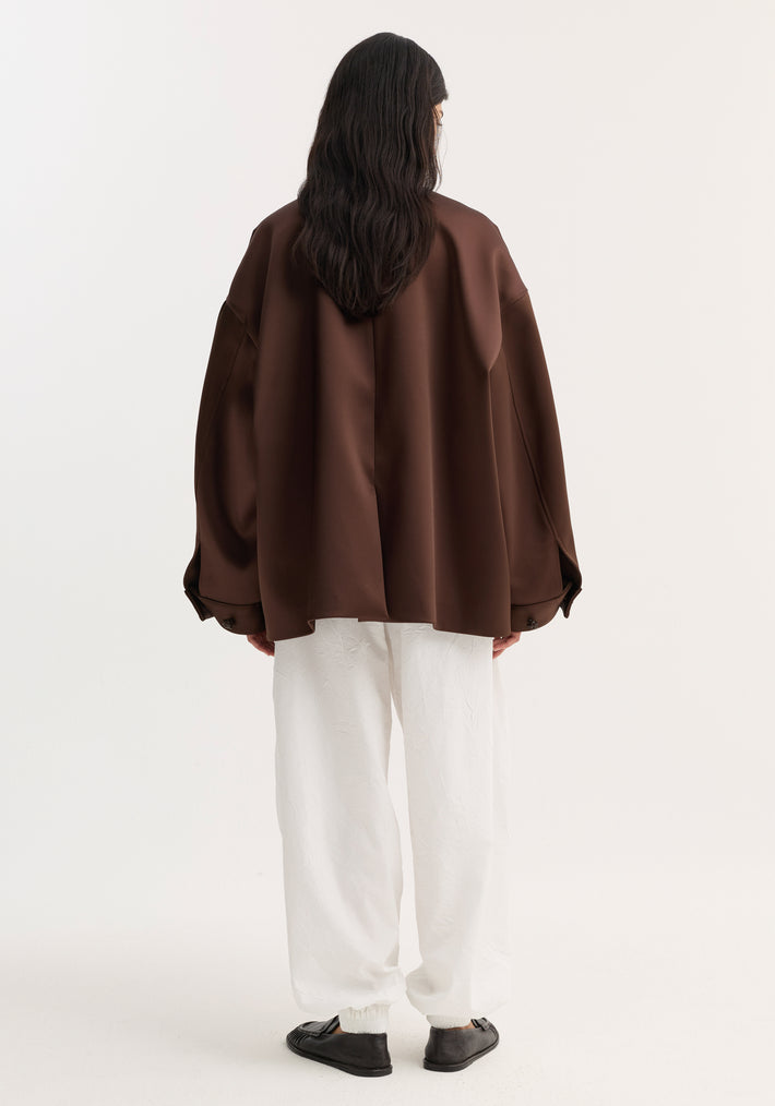 Back view of a model in an oversized Western heavy satin jacket with leather-knotted buttons and chest welt pockets, paired with white tapered trousers.